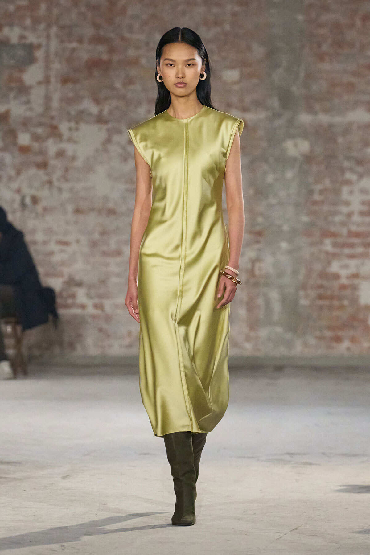 Model walks a fashion show in a gold satin sleeveless midi dress with cap shoulders and knee-high boots in front of a brick backdrop.
