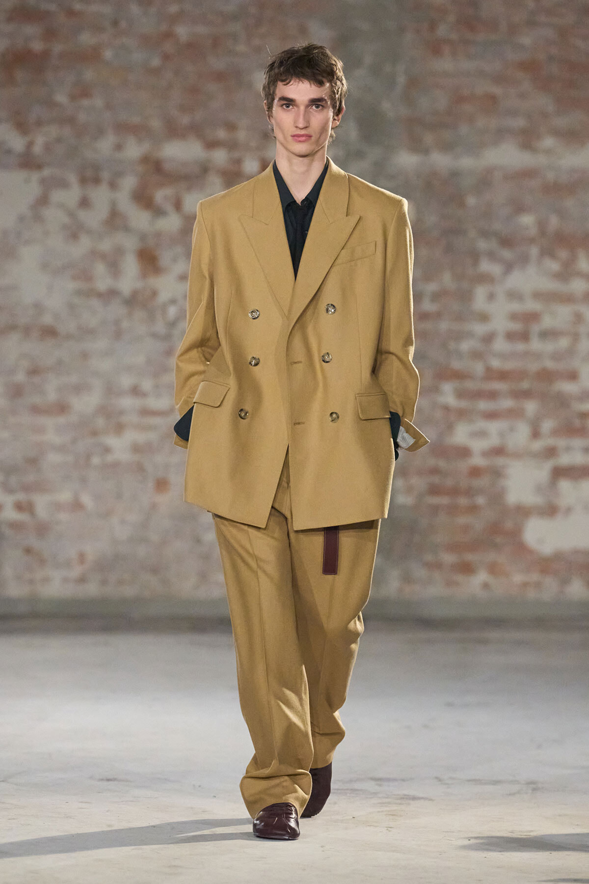 Male model on a runway wearing a beige double-breasted suit with a black shirt.