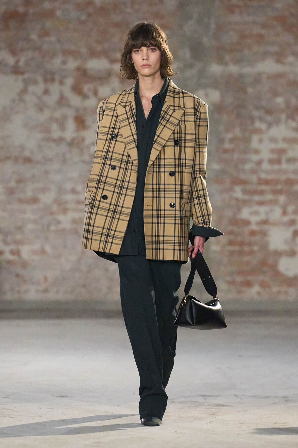 Runway model in a beige plaid double-breasted blazer over black shirt and trousers, carrying a black handbag against an exposed brick wall.