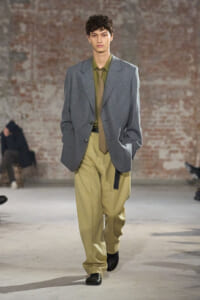 Male model on a runway wearing an oversized gray blazer, olive shirt, tan tie, and beige wide-leg pants against a brick wall backdrop.