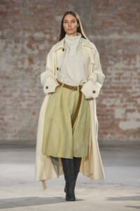 Model walks a runway in a long cream trench over a light beige top and olive-green midi skirt with a brown belt and black knee-high boots against a brick wall.