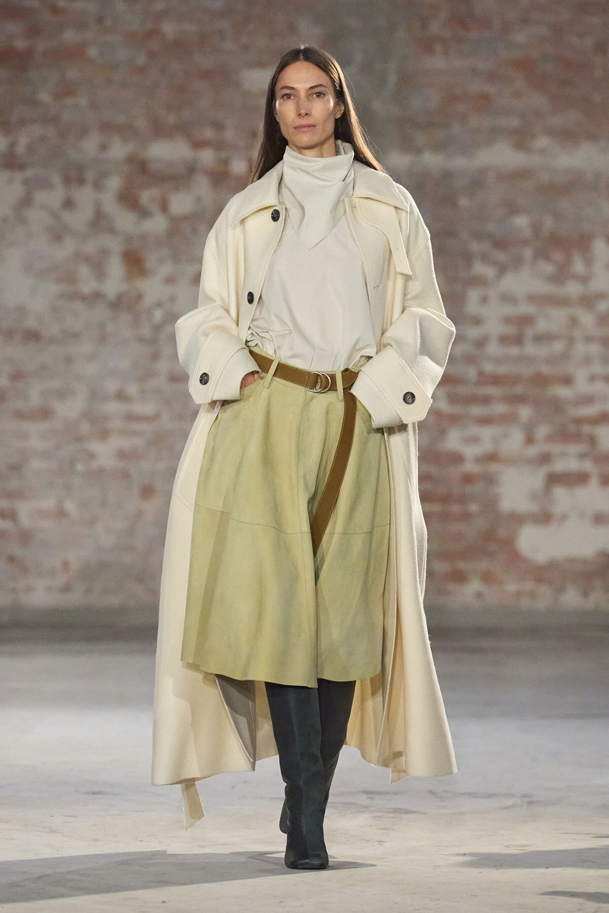 Model walks a runway in a long cream trench over a light beige top and olive-green midi skirt with a brown belt and black knee-high boots against a brick wall.