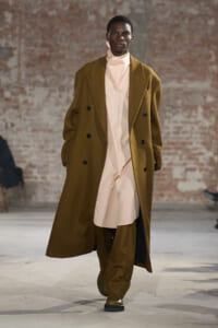 Male model walks a fashion runway in a long camel overcoat over a beige high-neck outfit, with matching trousers.
