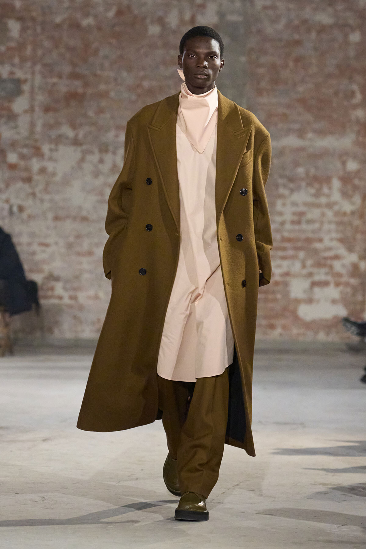 Male model walks a fashion runway in a long camel overcoat over a beige high-neck outfit, with matching trousers.