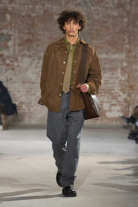 Male model walking on runway in a brown suede jacket over an olive shirt and tie, gray wide-leg trousers, black shoes, and a brown shoulder bag.