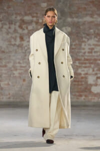 Model walking on a runway in a long white double-breasted coat over a black turtleneck, brick wall backdrop.