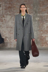 Model walking a runway in a long gray double-breasted coat, floral shirt, black tie, black trousers, and a brown leather tote bag at side.