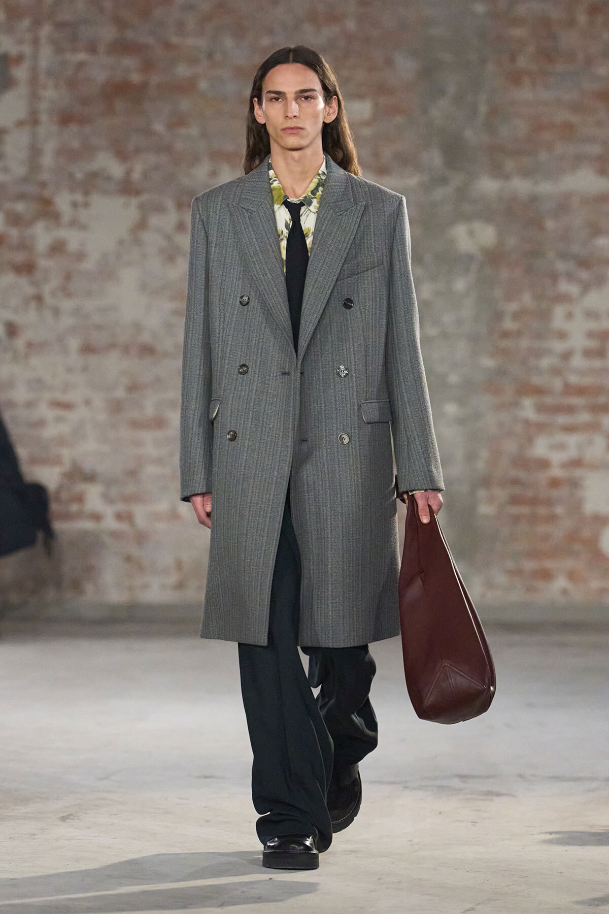 Model walking a runway in a long gray double-breasted coat, floral shirt, black tie, black trousers, and a brown leather tote bag at side.