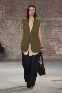 Model on a runway wearing an olive green sleeveless vest over a beige button-down shirt, paired with black wide-leg pants and a brown bag.