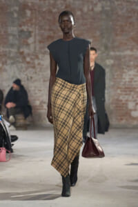 Model walking a runway in a dark sleeveless top and beige plaid wrap skirt, carrying a burgundy handbag.