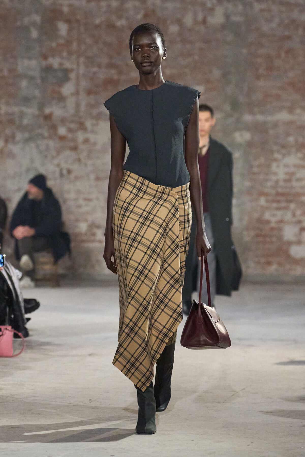 Model walking a runway in a dark sleeveless top and beige plaid wrap skirt, carrying a burgundy handbag.