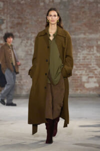 Model walking a runway in an olive blouse, oversized brown coat, brown suede pants, and burgundy boots against a brick wall backdrop.