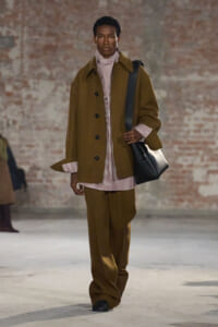 Male model on a runway wearing an oversized brown coat over a striped burgundy-and-white shirt and matching wide-leg pants, carrying a black shoulder bag