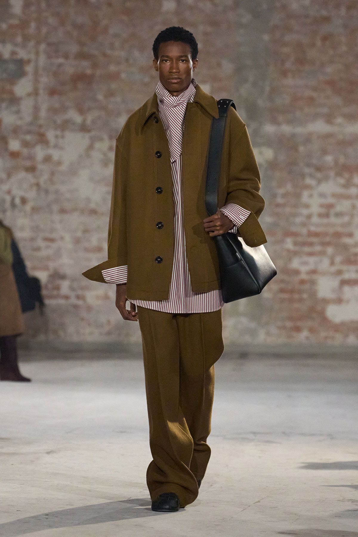 Male model on a runway wearing an oversized brown coat over a striped burgundy-and-white shirt and matching wide-leg pants, carrying a black shoulder bag