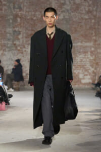 Male model on a fashion runway wearing a dark double‑breasted overcoat over a burgundy sweater and gray trousers, carrying a black bag.