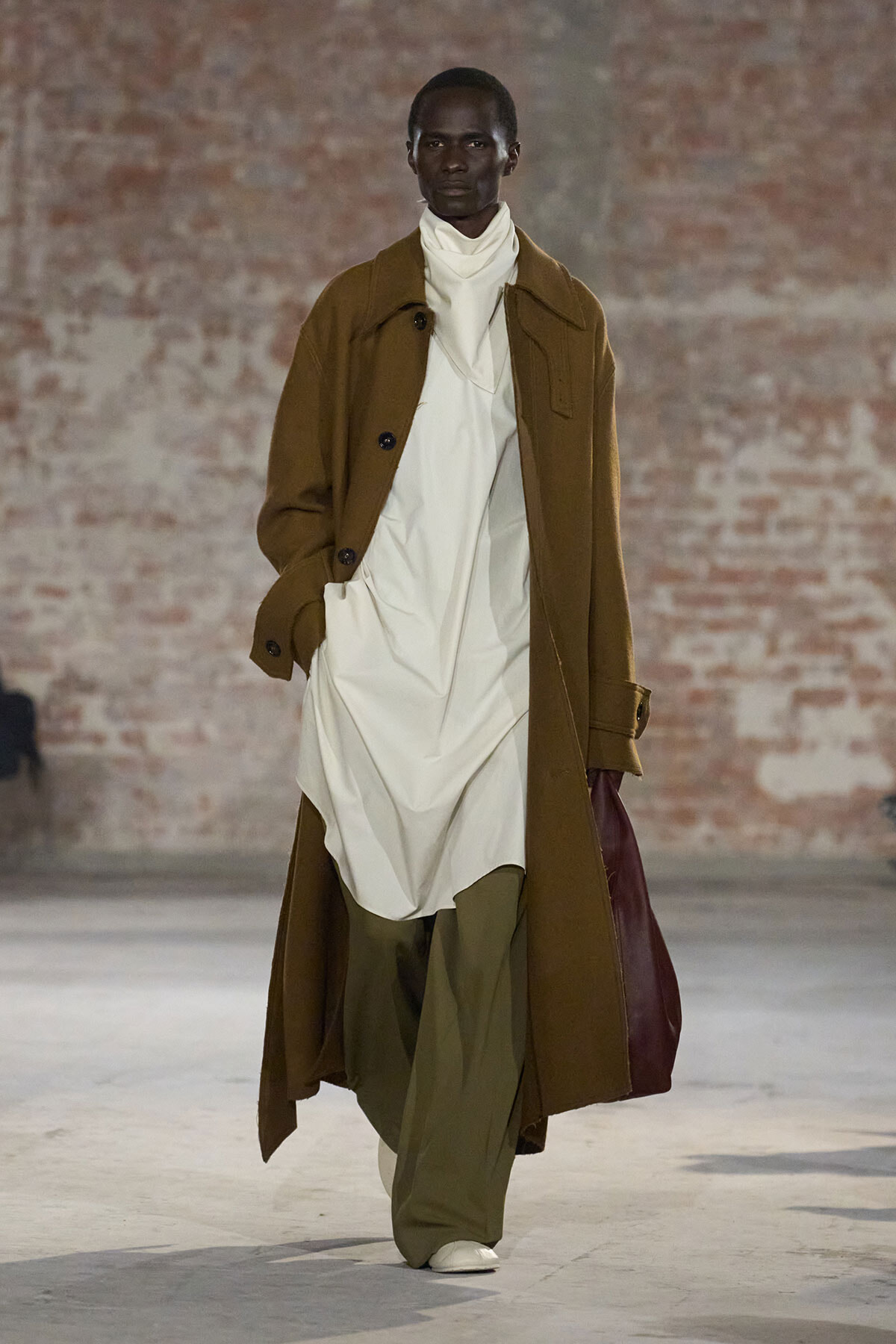 Male model walking on runway in a camel overcoat over a white layered outfit and olive wide-leg pants; brick wall backdrop.