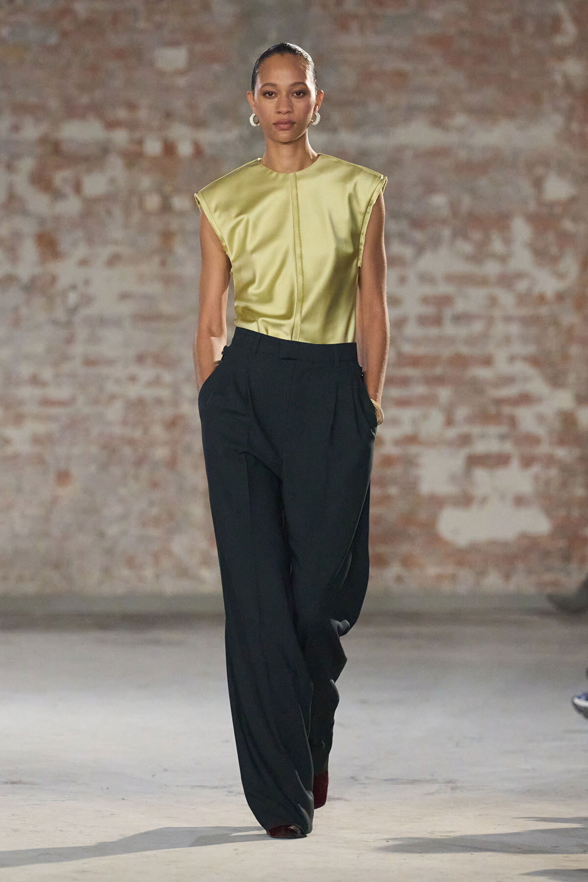 Runway model wearing a metallic gold sleeveless top and wide-leg black trousers, hands in pockets, in front of an exposed brick wall.