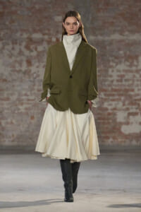 Model walking a runway in an olive blazer, cream turtleneck, and a full cream pleated skirt with black knee-high boots against a brick wall backdrop.