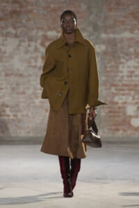 Model walks a runway in an oversized olive-brown coat, brown turtleneck, and a knee-length skirt, paired with burgundy boots and a brown handbag.
