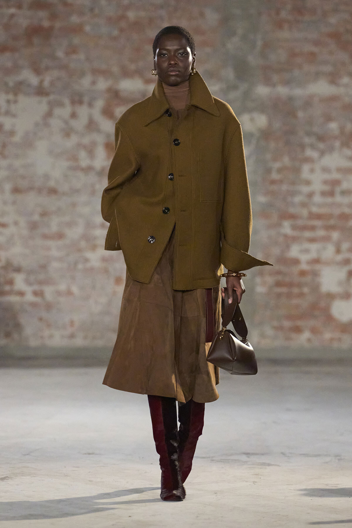 Model walks a runway in an oversized olive-brown coat, brown turtleneck, and a knee-length skirt, paired with burgundy boots and a brown handbag.