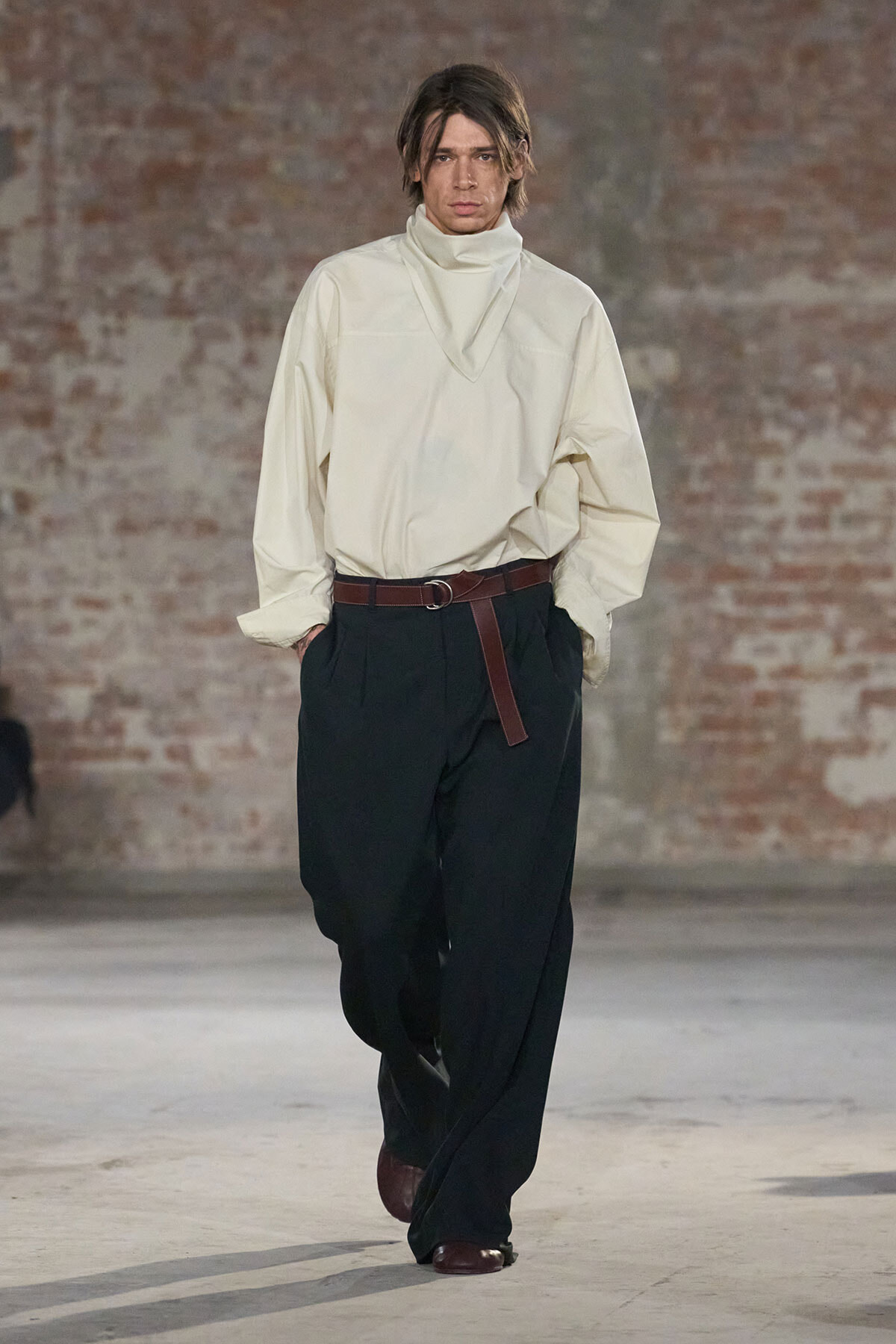 Male model walks a fashion runway in a loose cream turtleneck top with a high neck, black wide-leg trousers, and a brown belt; brick wall backdrop.