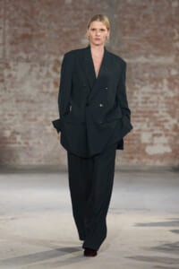Model walks a runway in an oversized black double-breasted blazer and wide-leg trousers, with a brick-wall backdrop.