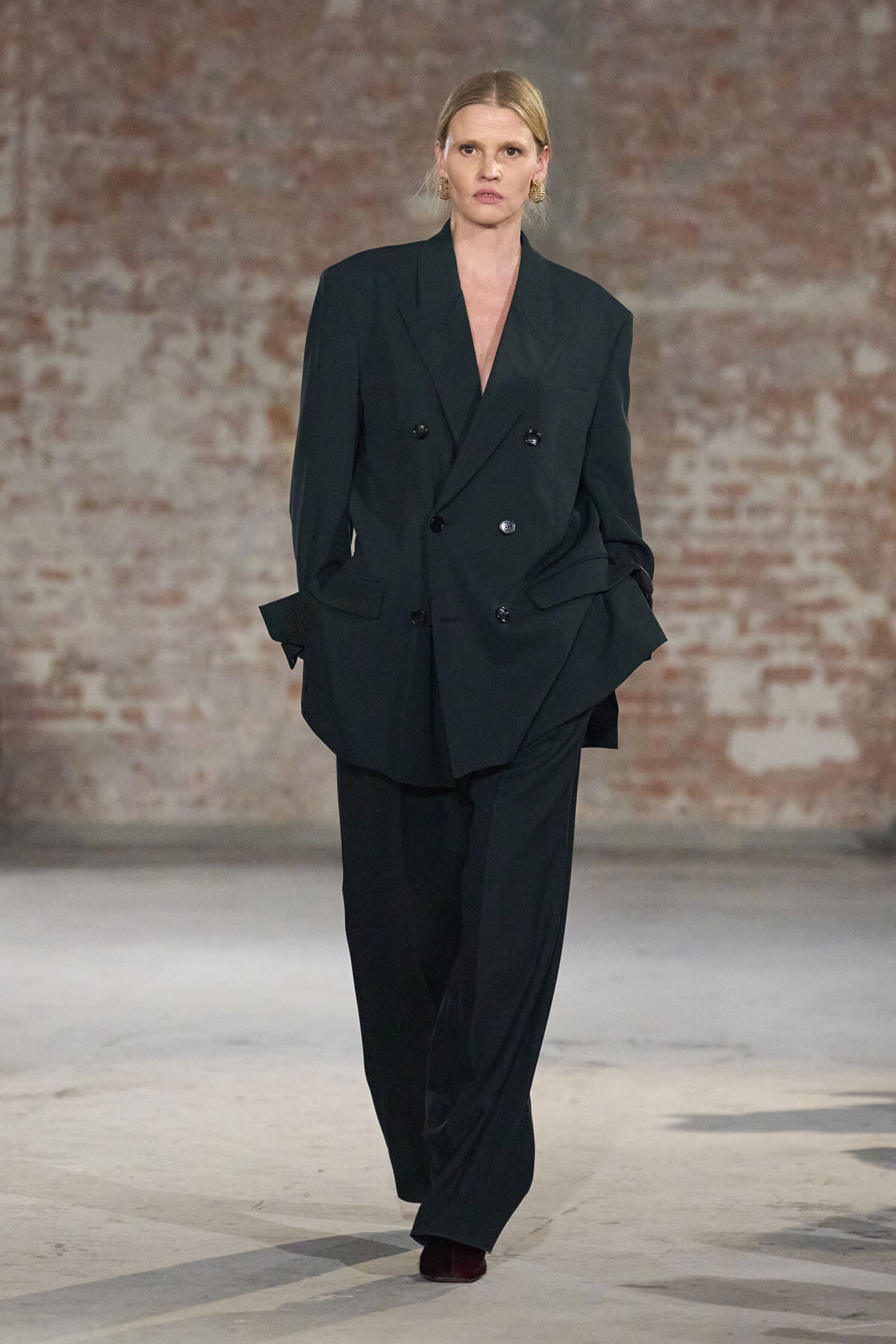 Model walks a runway in an oversized black double-breasted blazer and wide-leg trousers, with a brick-wall backdrop.
