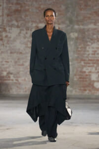 Model wears an oversized black double-breasted blazer with a long, layered skirt, walking a runway while carrying a metallic clutch.