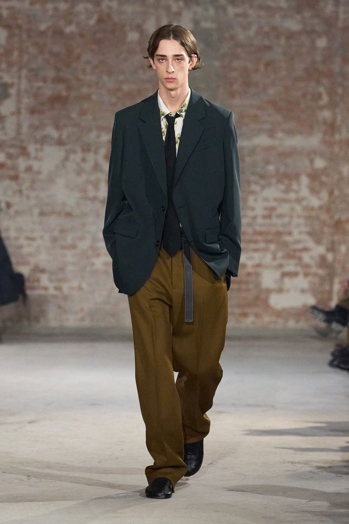 Model on a fashion runway wearing a dark blazer, floral shirt, black tie, and wide brown trousers in front of a brick wall.