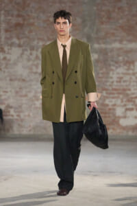 Male model walks a runway wearing an oversized olive double-breasted blazer, beige shirt, brown tie, dark trousers, and a black bag in hand.