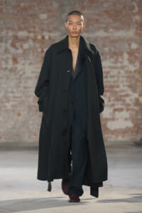 Male model walking a fashion runway in a long dark overcoat over black clothing, brick wall backdrop behind.