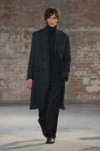 Male model walks a runway in a dark double-breasted overcoat and black turtleneck, brick backdrop behind him.