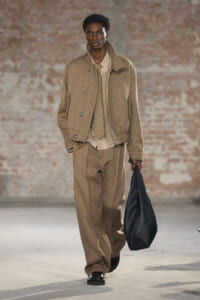 Male model walking a runway in a beige corduroy suit with oversized jacket and wide trousers, carrying a black bag.
