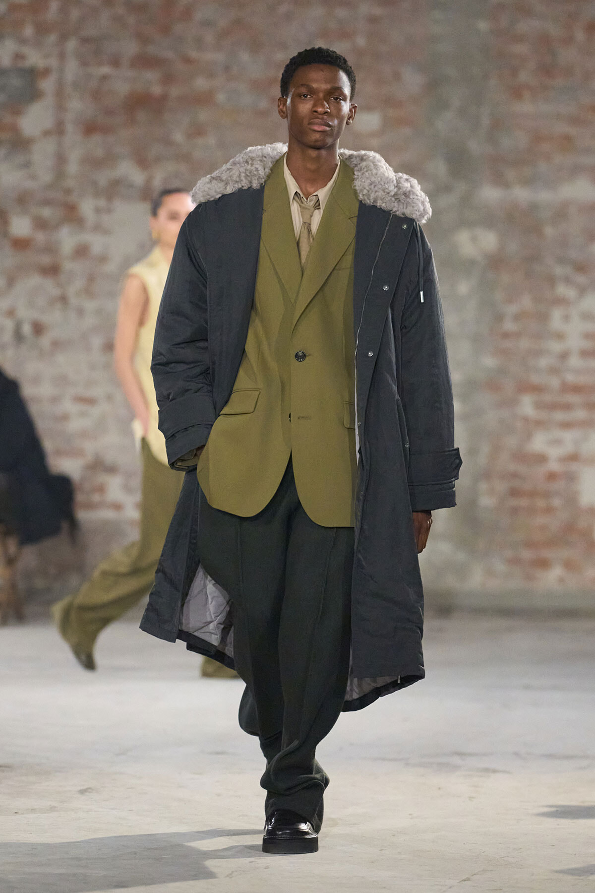 Male model walking a fashion runway in an olive suit under a long black parka with a fur-lined collar, industrial brick backdrop.