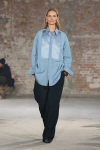 Model walks a runway in a light blue oversized shirt with satin front panels and black wide-leg trousers, brick wall backdrop behind.