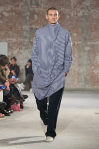 Male model walking a fashion runway in a loose, blue-and-white striped tunic with a matching scarf, hands in pockets, audience seated along a brick-wall backdrop.