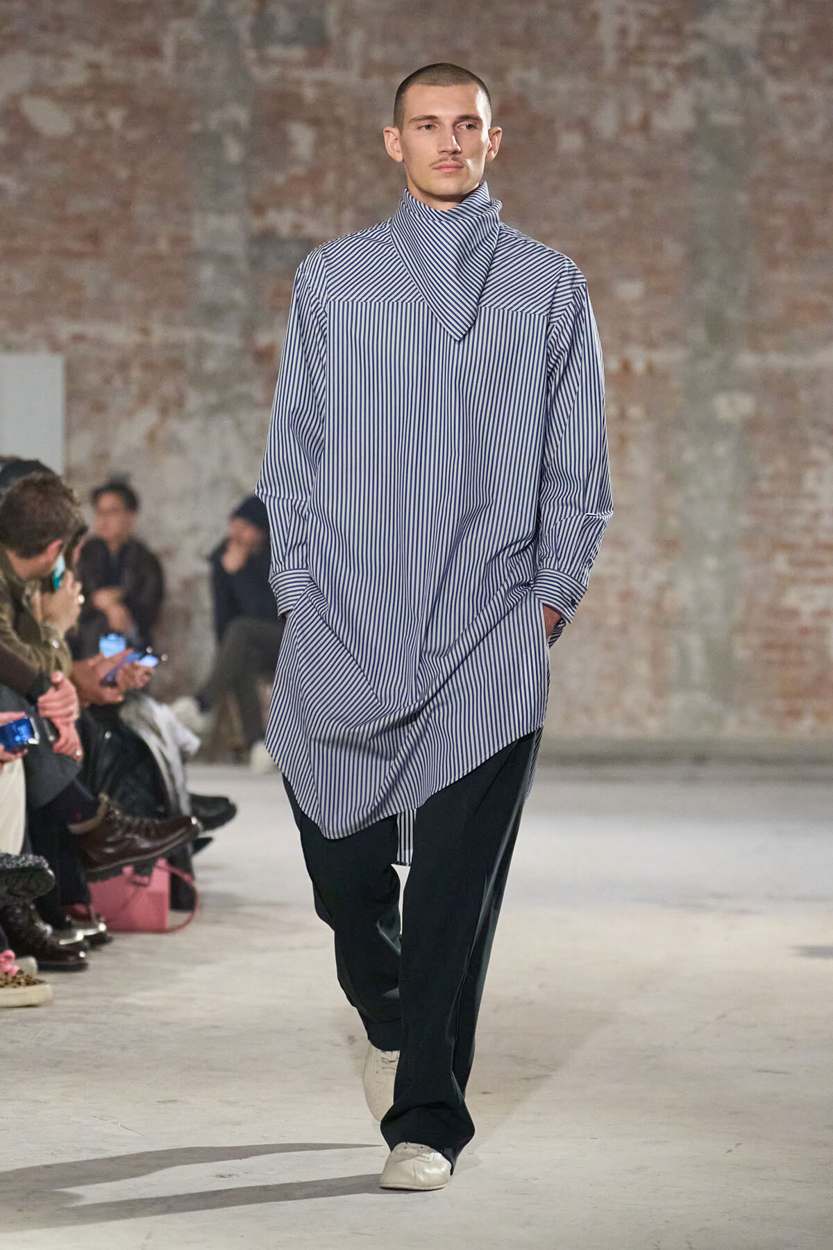 Male model walking a fashion runway in a loose, blue-and-white striped tunic with a matching scarf, hands in pockets, audience seated along a brick-wall backdrop.