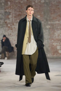 Male model walking a fashion runway in a long black coat over layered neutrals, olive trousers, and a beige shirt with tan tie against a brick wall backdrop.