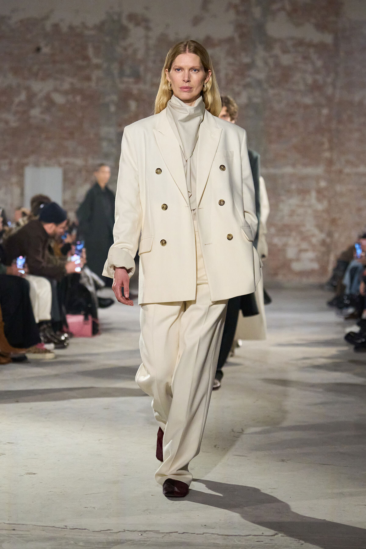 Female model walks a fashion show runway in a cream oversized double-breasted suit with a turtleneck.