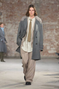 Male model walks the runway in an oversized gray plaid coat with a fur collar, cream shirt, tan tie, and baggy beige trousers, against a brick wall backdrop.