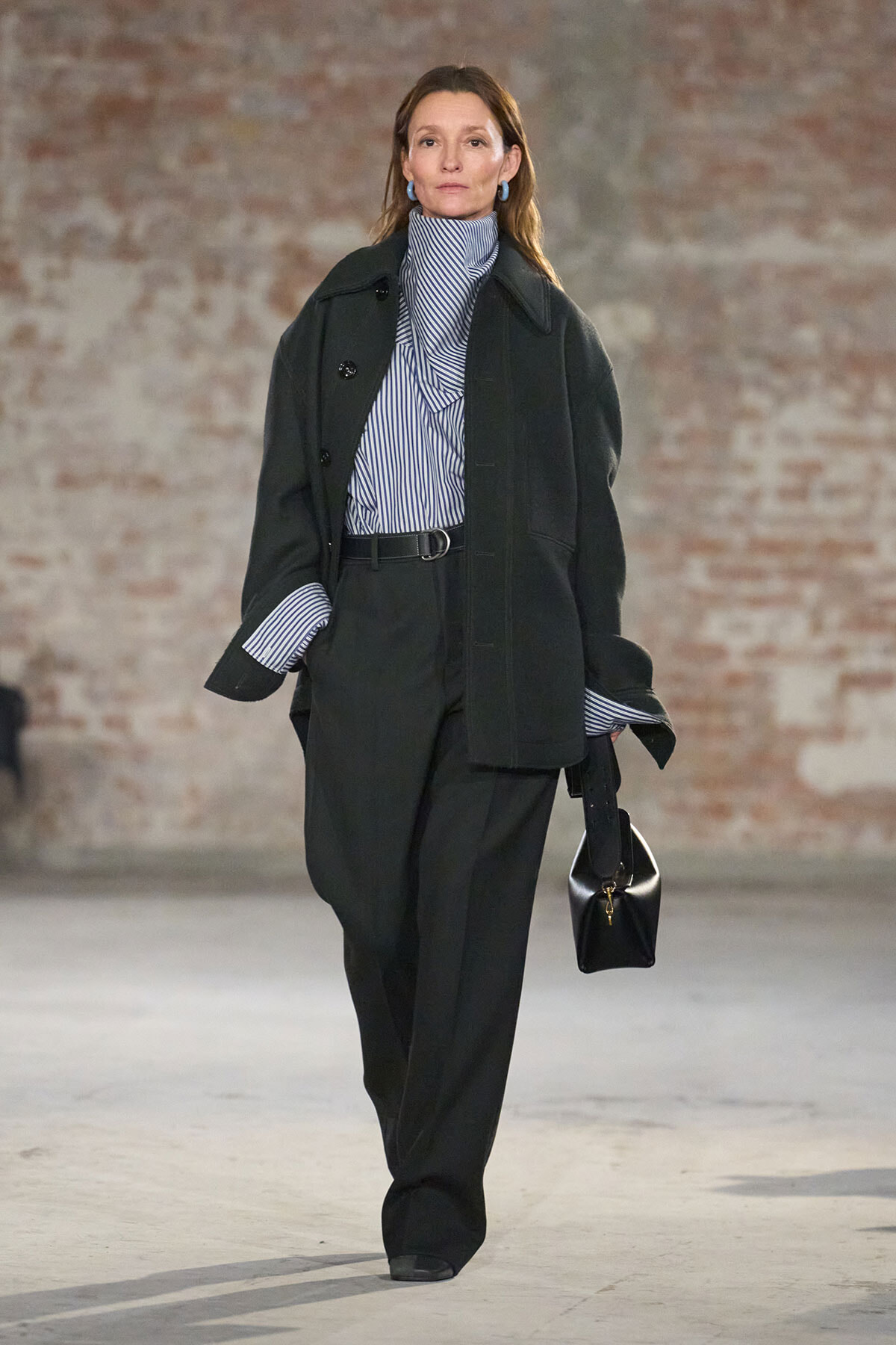 Female model wearing a dark oversized coat over a striped shirt, wide-leg pants, and a black handbag on a runway.