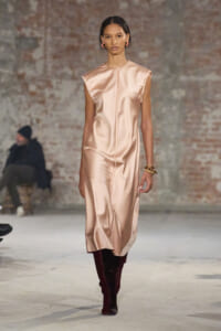 Model walking the runway in a peach satin sleeveless midi dress with a center seam, paired with burgundy knee-high boots and gold bracelets.