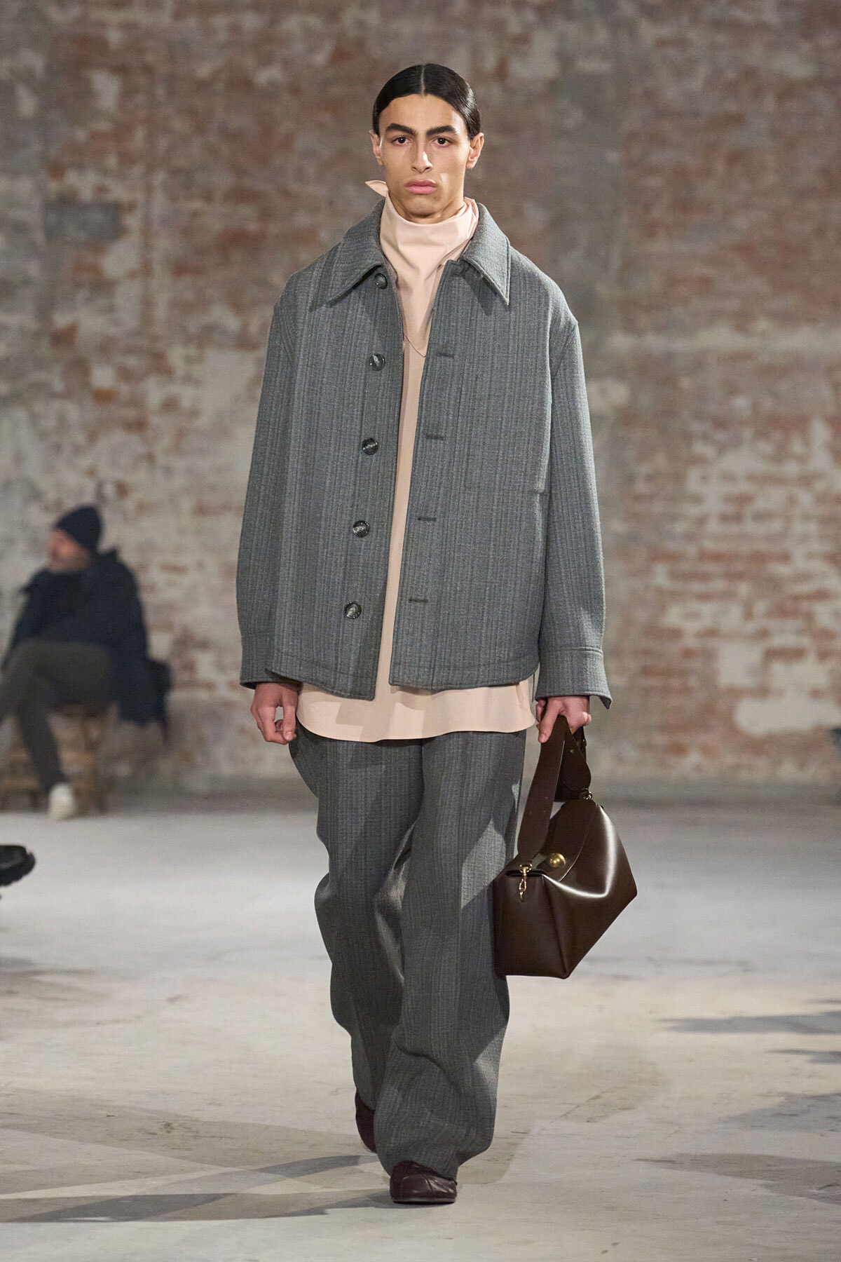 Model walks a runway wearing a loose gray pinstripe suit with a beige layered top and matching wide-leg pants, carrying a brown leather duffel bag.