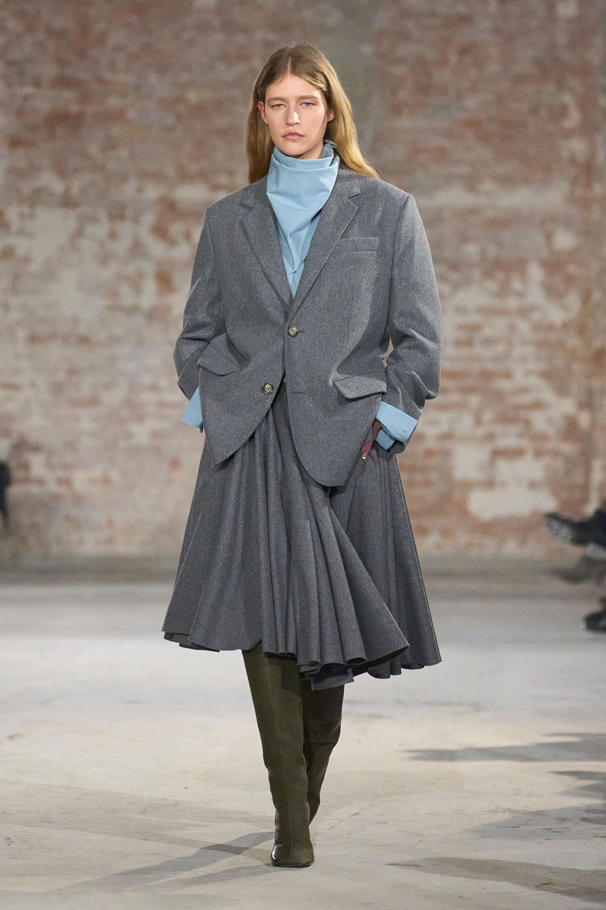 Model on a runway wearing an oversized gray blazer, blue turtleneck, and matching pleated gray skirt with tall brown boots a brick backdrop behind.