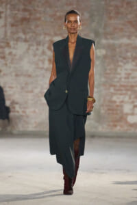 Model walks the runway in a dark green sleeveless blazer and matching long skirt, with maroon boots and gold jewelry against a brick wall.