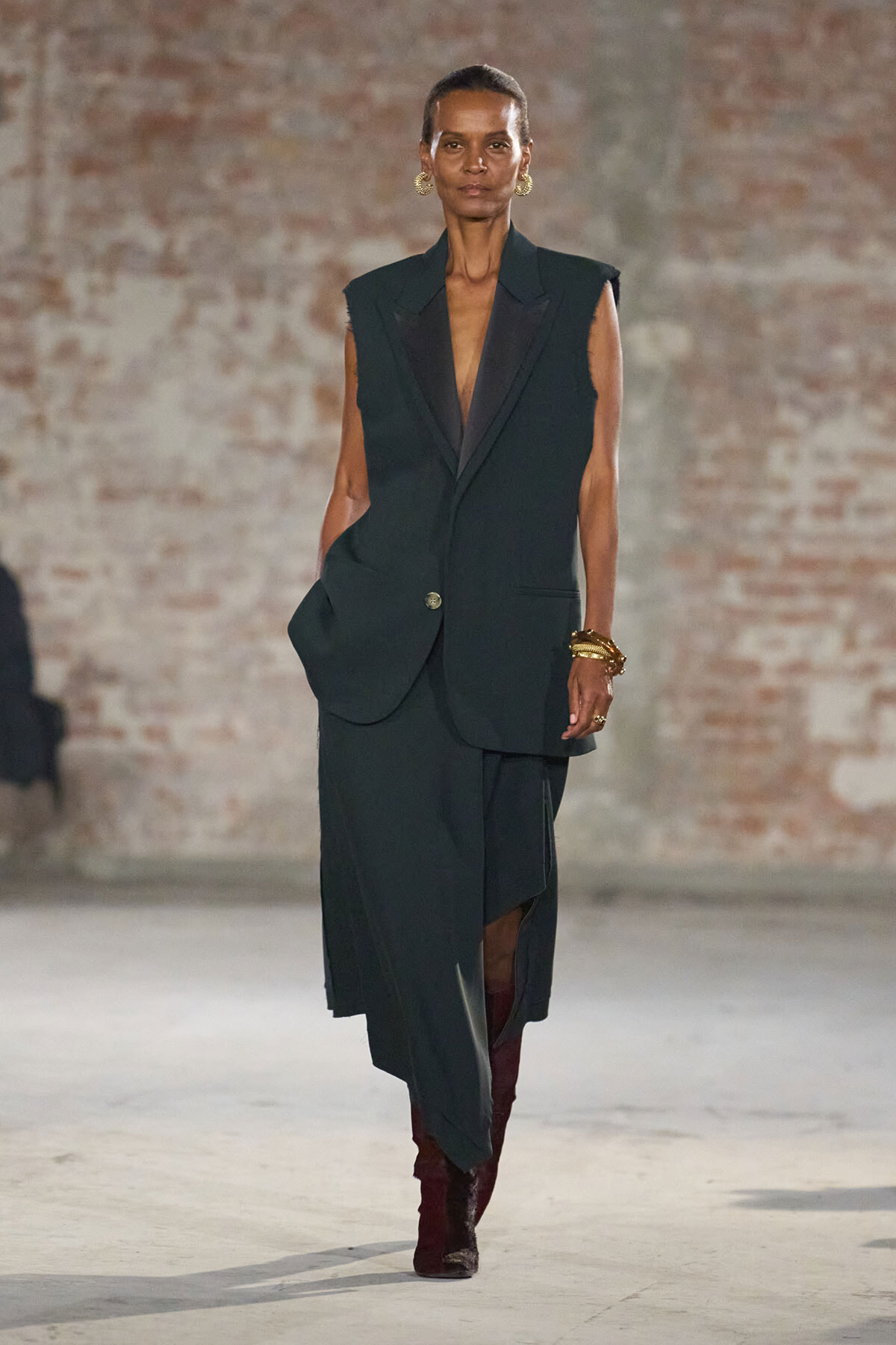 Model walks the runway in a dark green sleeveless blazer and matching long skirt, with maroon boots and gold jewelry against a brick wall.