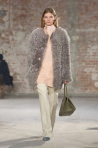 Model walks runway in a large gray faux-fur coat over a pale pink tunic, cream trousers, and a taupe handbag.