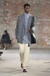 Male model on a runway wearing a gray double-breasted blazer over a blue-and-white striped shirt, cream wide-legged trousers, burgundy shoes, and a black shoulder bag.