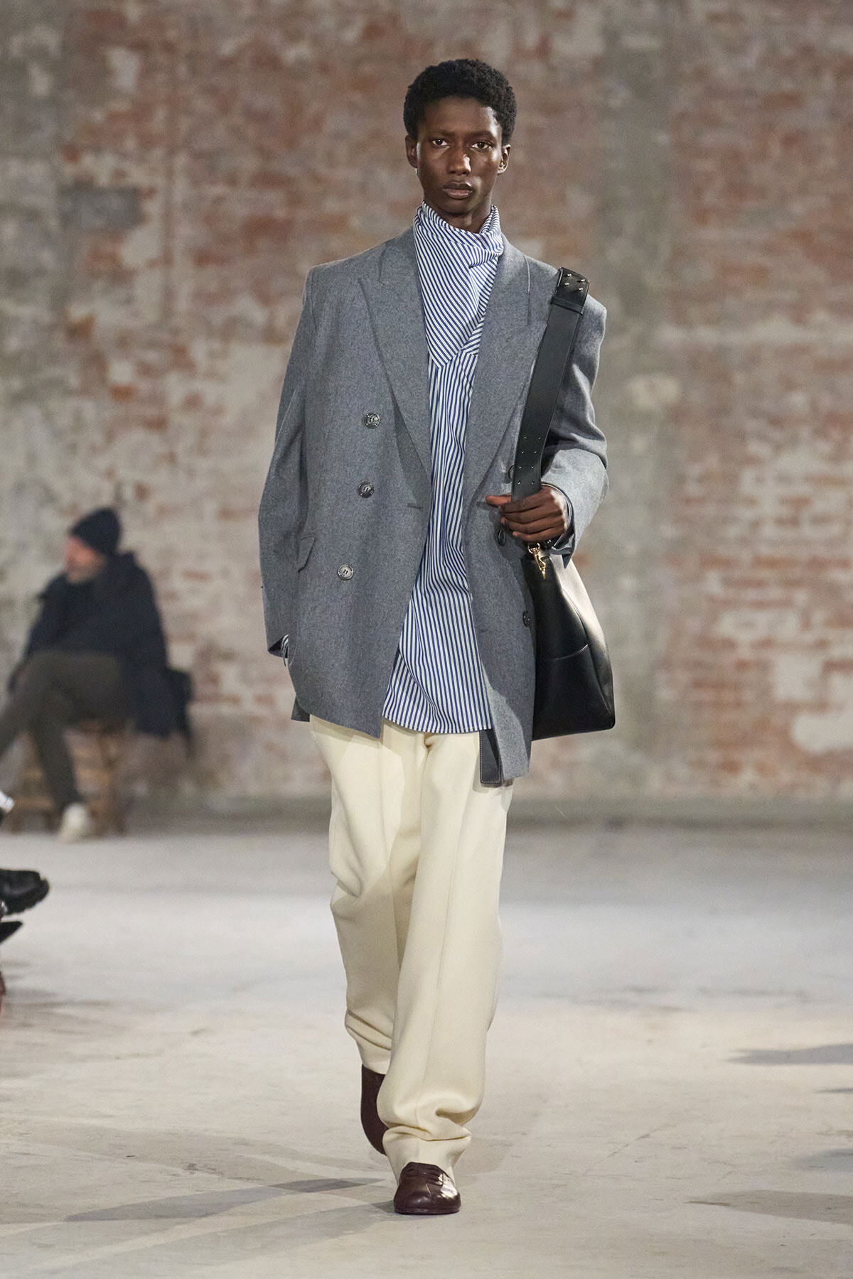 Male model on a runway wearing a gray double-breasted blazer over a blue-and-white striped shirt, cream wide-legged trousers, burgundy shoes, and a black shoulder bag.