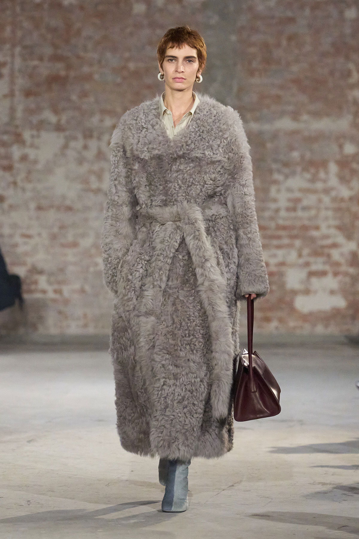 Model on a runway wearing a long gray faux‑fur coat belted at the waist, with a pale shirt, light blue boots, and a burgundy handbag against a brick wall backdrop.